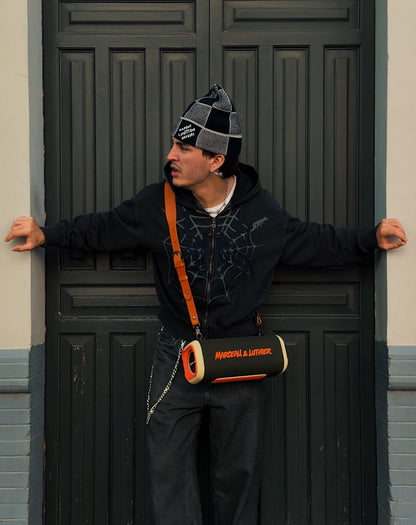 A model posing in front of a dark paneled door wearing a checkered beanie and a black zip-up hoodie. He is wearing the Nocturne speaker across his chest using a long leather shoulder strap.