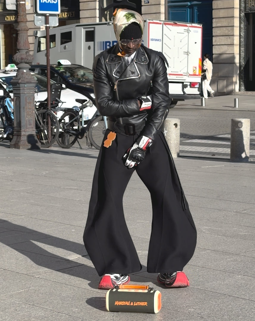 A model standing on a sunny sidewalk wearing a black leather biker jacket and oversized wide-leg trousers. The Nocturne speaker by Marceau & Luthier is placed on the ground at his feet.