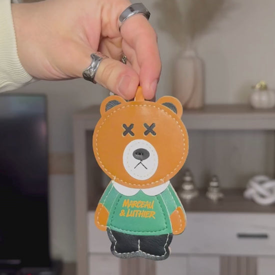A video showcasing accessories for the Nocturne portable speaker. It begins with a person’s hand, wearing silver rings, holding up a brown leather bear charm with "Marceau & Luthier" printed on its green sweater. The video then shows an orange leather strap with the brand name embossed, followed by a close-up of a metal clasp and an embossed bear logo on the leather.